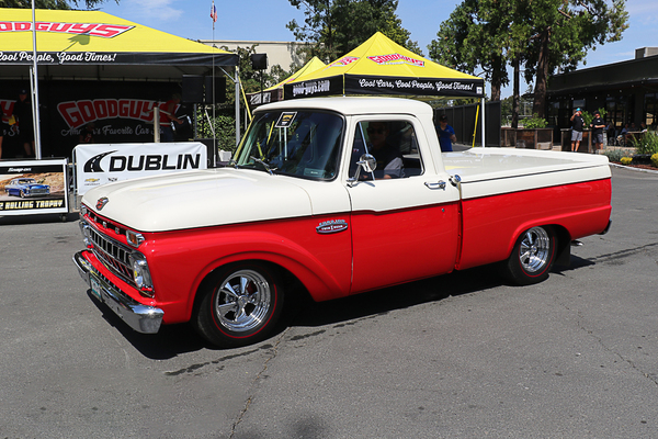 Front three-quarter view of a lowered two-tone red and white 1965–1966 Ford F-100 Twin I-Beam pickup truck driving past the Goodguys tent at the Goodguys Pleasanton West Coast Nationals.