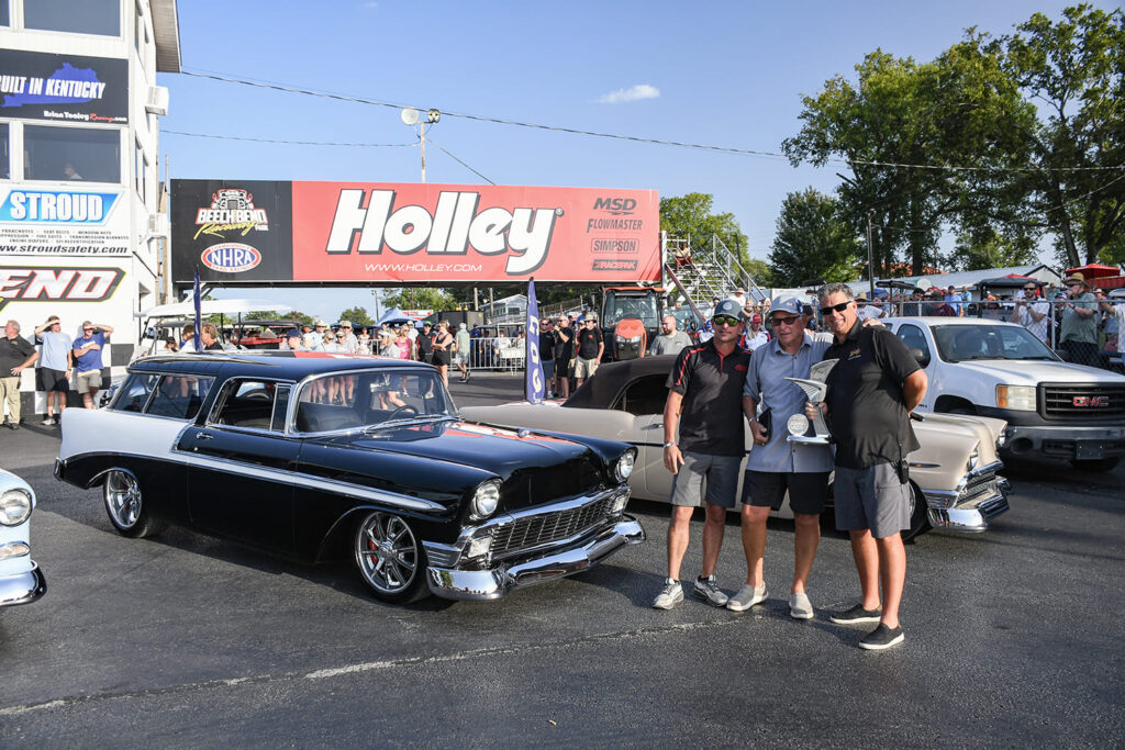 Car owners and builders posing with a trophy next to a black and white 1956 Chevrolet Nomad in front of the Holley tower.