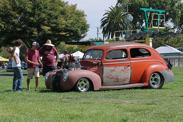 Side profile of an orange-red patina 1940 Ford two-door sedan street rod with exposed engine velocity stacks parked on the grass at the Goodguys Pleasanton West Coast Nationals.