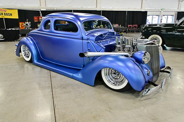 Front three-quarter view of a slammed satin matte blue 1935–1936 Ford five-window coupe street rod with an exposed multi-carburetor engine on display indoors at the Goodguys Pleasanton West Coast Nationals.