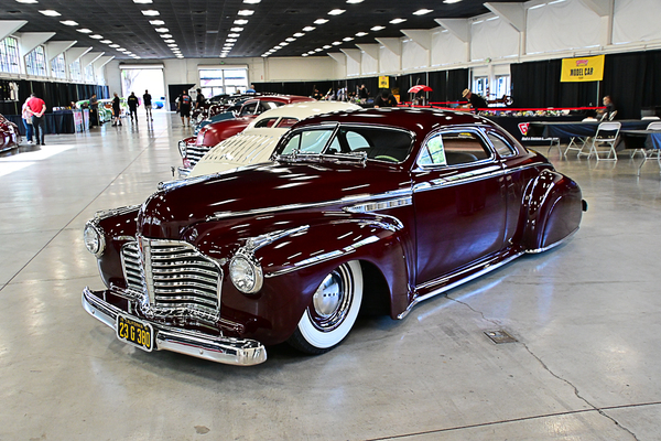 Front three-quarter view of a slammed deep burgundy chopped 1941 Buick custom coupe with wide whitewall tires on display indoors at the Goodguys Pleasanton West Coast Nationals.