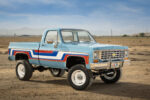 Rockford Smith’s “Spirit of ‘76” Scratch-Built Chevy K10 Front three-quarter view of a lifted 1976 Chevy K10 4x4 pickup truck painted light blue with red, white, and blue vintage side stripes, chrome bumper, and white off-road wheels in an open desert.