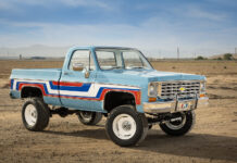 Front three-quarter view of a lifted 1976 Chevy K10 4x4 pickup truck painted light blue with red, white, and blue vintage side stripes, chrome bumper, and white off-road wheels in an open desert.