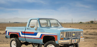 Front three-quarter view of a lifted 1976 Chevy K10 4x4 pickup truck painted light blue with red, white, and blue vintage side stripes, chrome bumper, and white off-road wheels in an open desert.