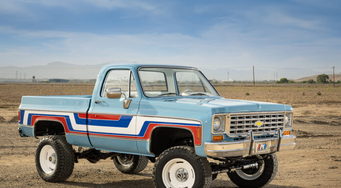 Front three-quarter view of a lifted 1976 Chevy K10 4x4 pickup truck painted light blue with red, white, and blue vintage side stripes, chrome bumper, and white off-road wheels in an open desert.