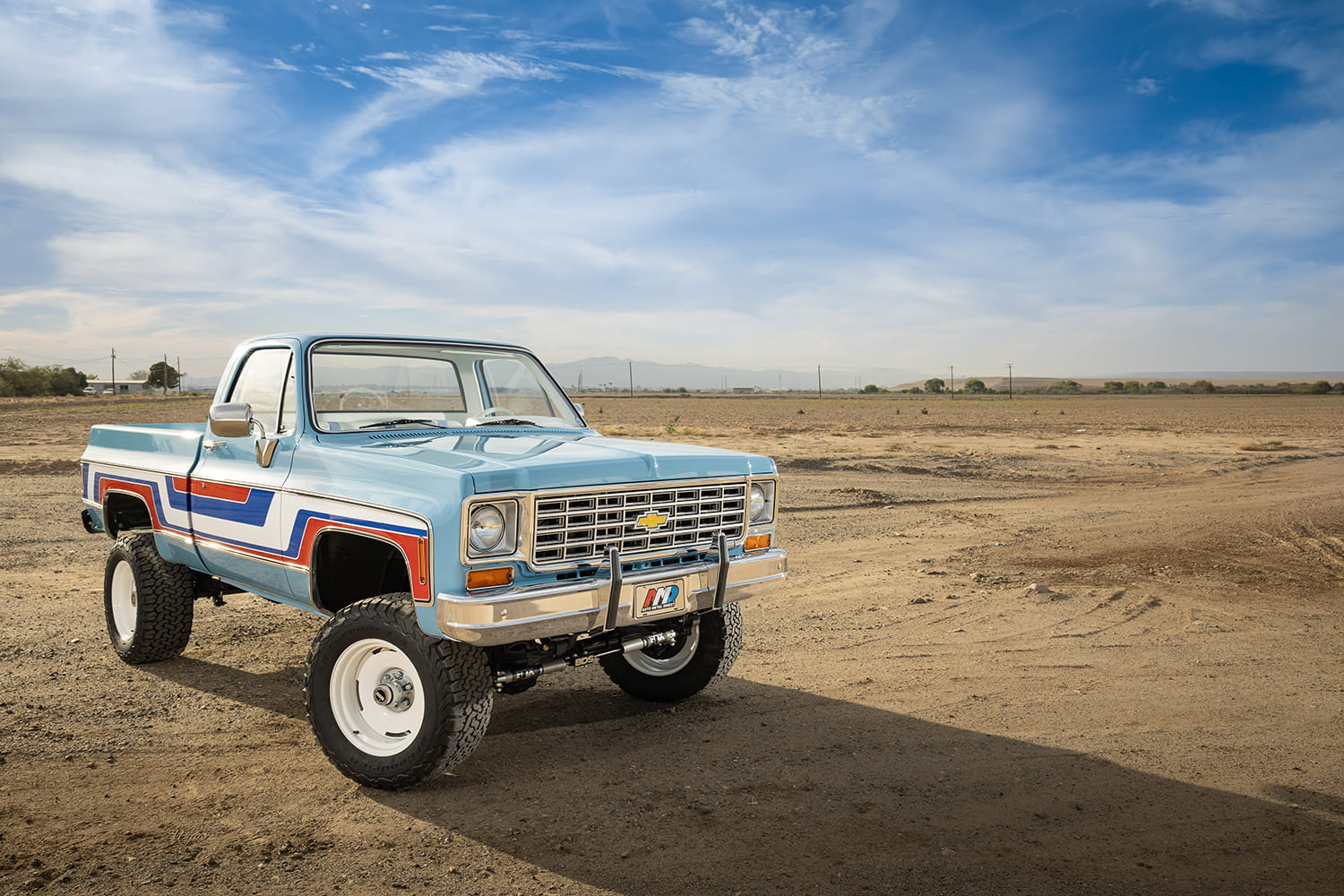 Wide front three-quarter view of a lifted light blue 1976 Chevy 4x4 pickup truck featuring vintage side stripes and white wheels parked in an open dirt desert under a blue sky.