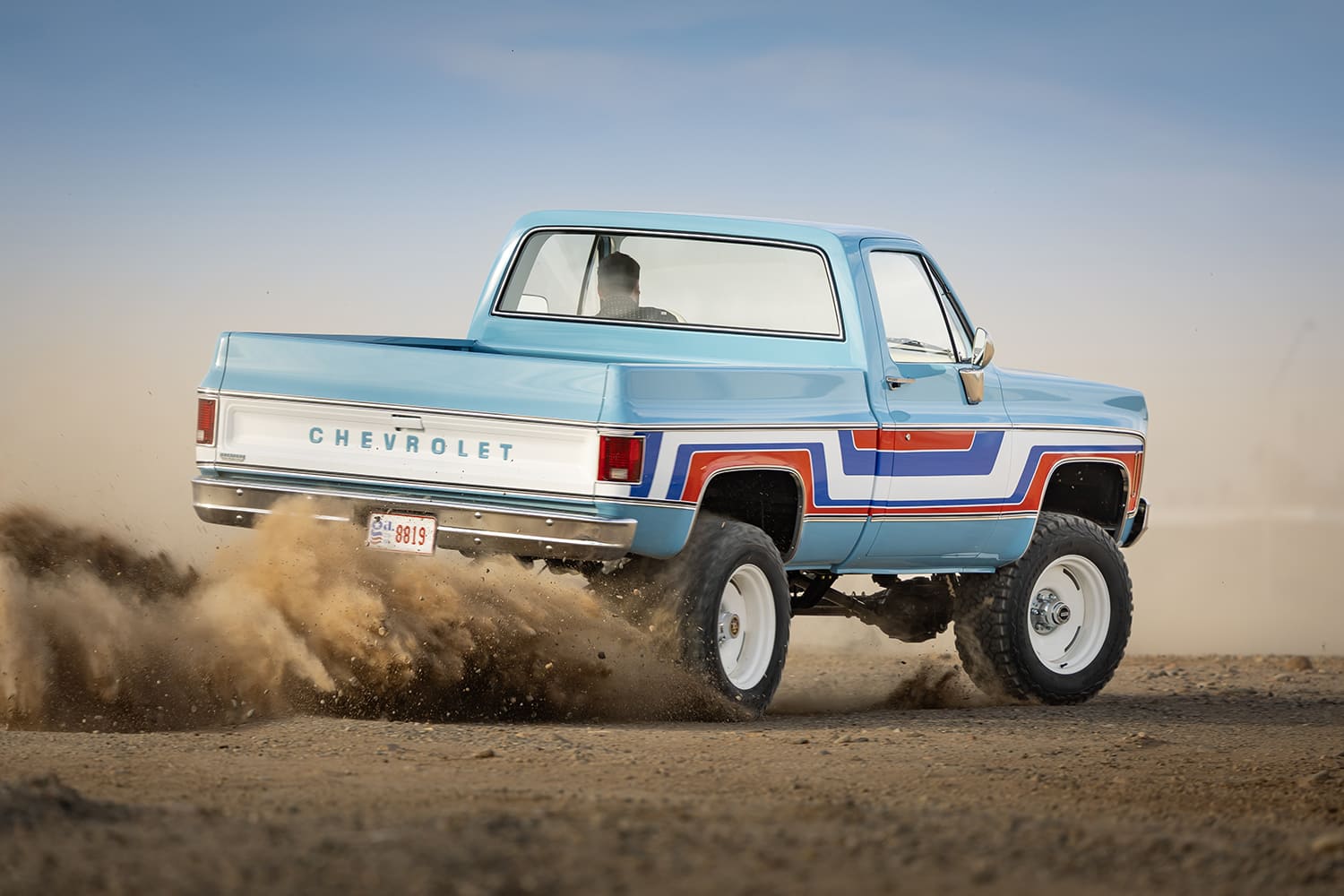 Dynamic rear three-quarter action shot of a lifted light blue 1976 Chevy 4x4 pickup truck kicking up a large cloud of dirt while driving off-road in the desert.