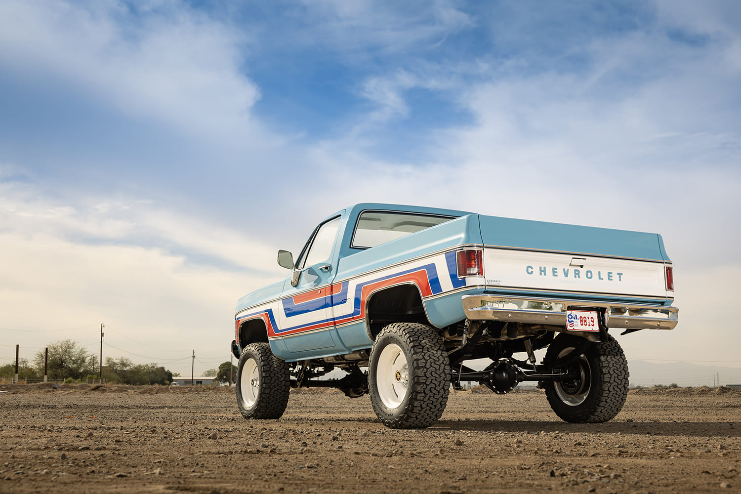 Low-angle rear three-quarter view of a lifted 1976 Chevy 4x4 pickup truck featuring light blue paint, bold red and blue body stripes, a white Chevrolet tailgate, and all-terrain tires on white steel wheels in the desert.