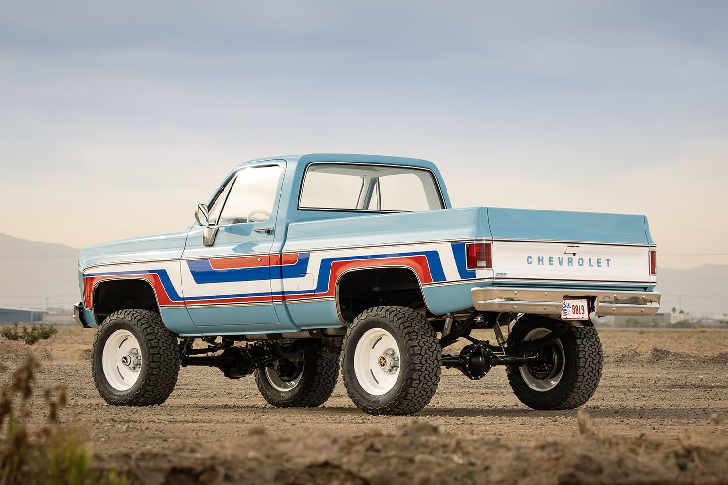 Rear three-quarter view of a lifted light blue 1976 Chevy 4x4 pickup truck parked in the desert, featuring bold retro side stripes, a white Chevrolet tailgate, and all-terrain tires on white wheels