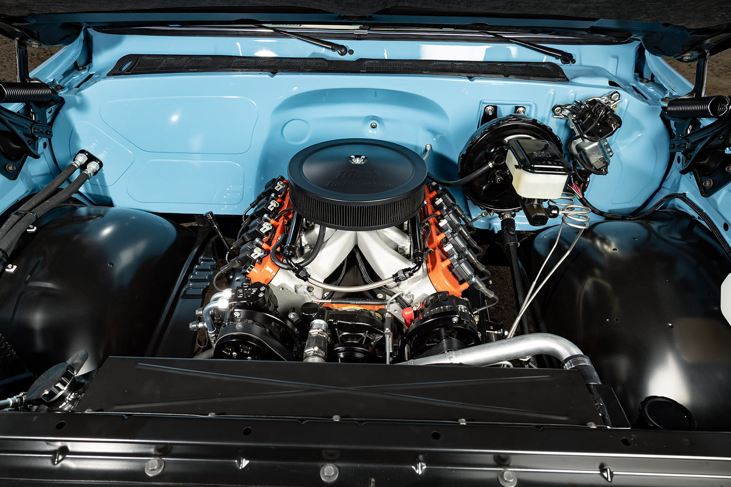 Top-down view of a restored 1976 Chevy pickup engine bay with color-matched light blue firewall, FiTech EFI intake, LS engine with orange valve covers, and black billet serpentine drive.