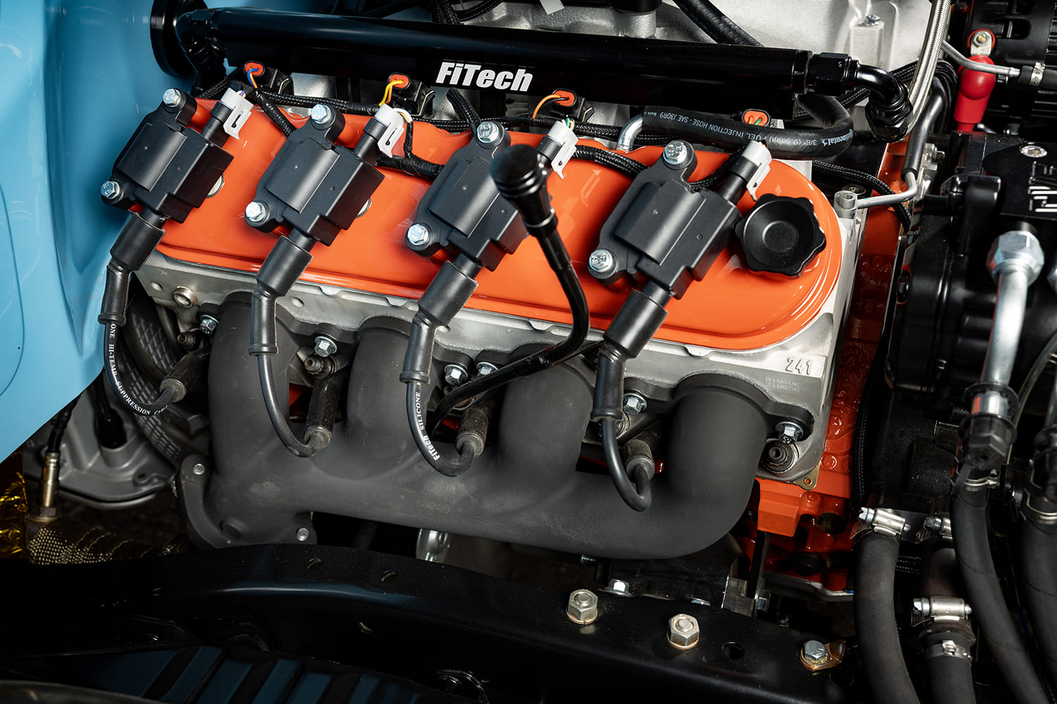 Close-up detail of a Chevy V8 engine cylinder head featuring Chevy orange valve covers with coil-on-plug ignition, FiTech fuel rail, and black exhaust headers.