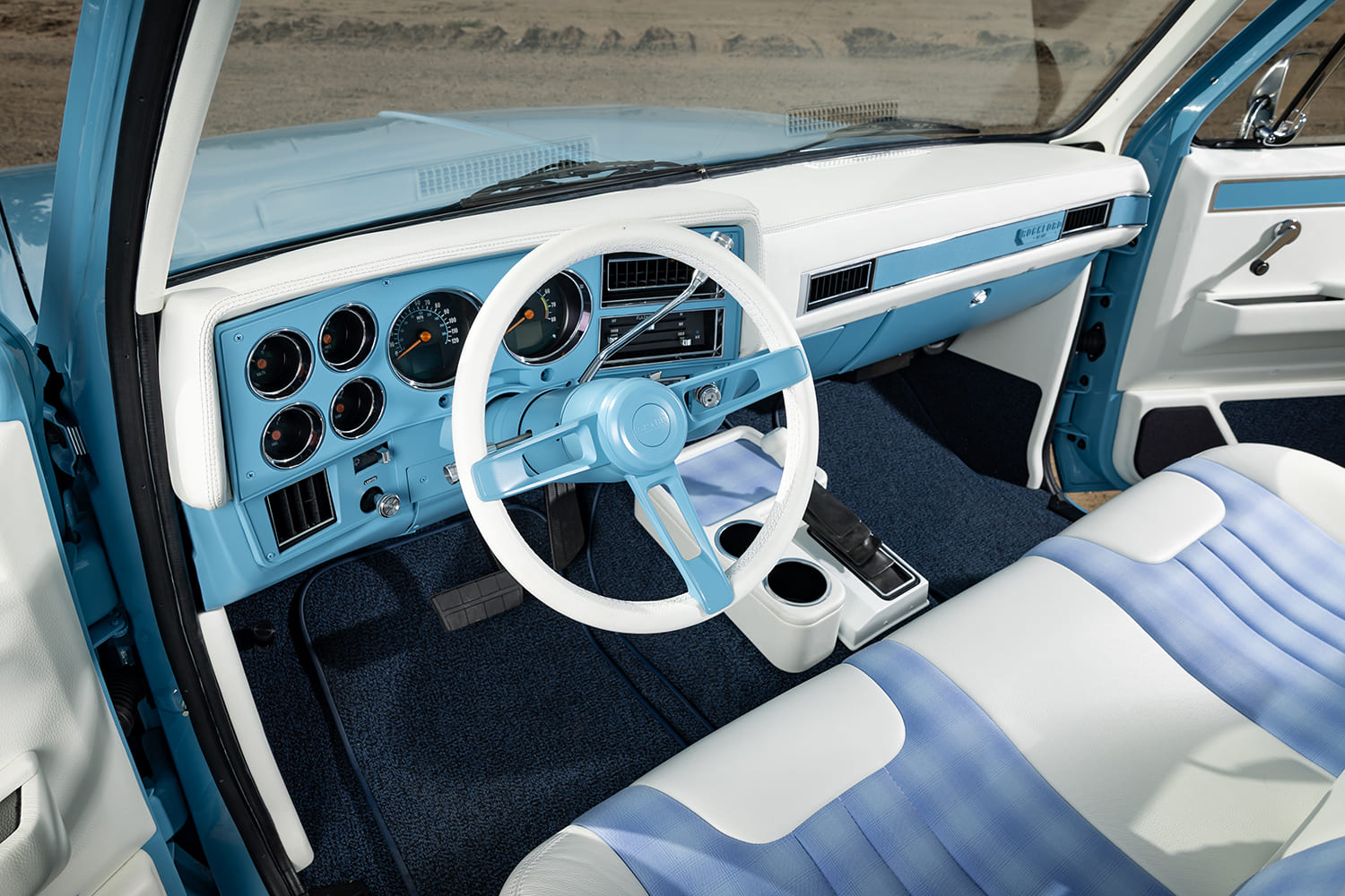 Custom two-tone interior of a 1976 Chevy pickup truck featuring a white and light blue steering wheel, modern digital gauges, stitched dash pad, and custom blue plaid bench seating.