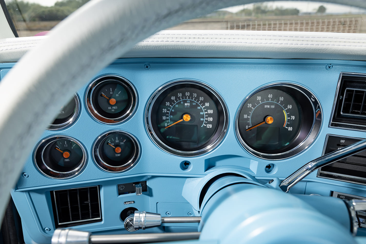 Close-up of the light blue dashboard instrument cluster in a 1976 Chevy pickup truck, featuring custom analog-style gauges with orange needles, chrome bezel rings, and a white steering wheel rim in the foreground.