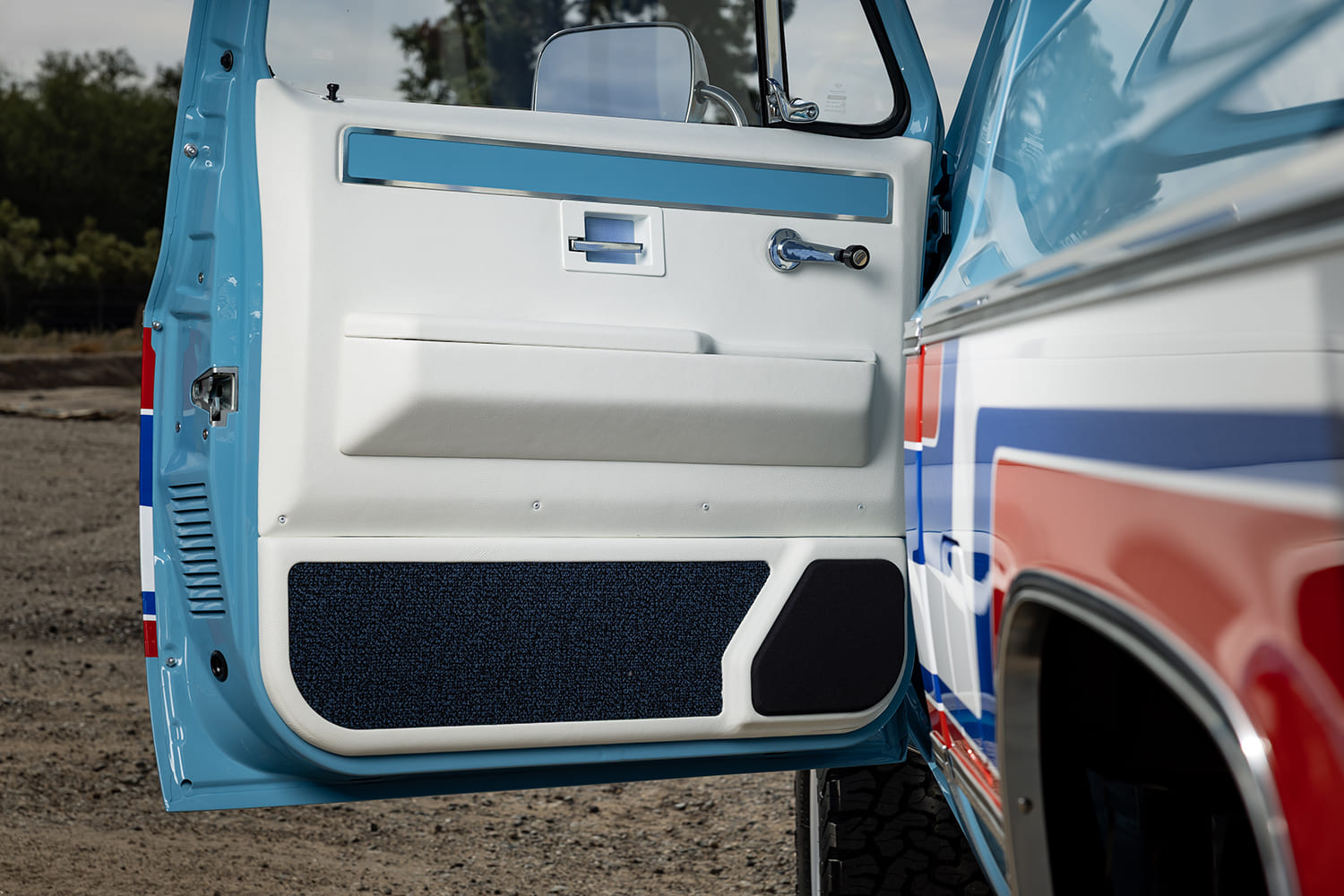 Detailed view of the open driver door on a 1976 Chevy pickup truck featuring a custom white upholstered door panel, blue carpet lower trim, integrated speaker grille, and chrome handle.