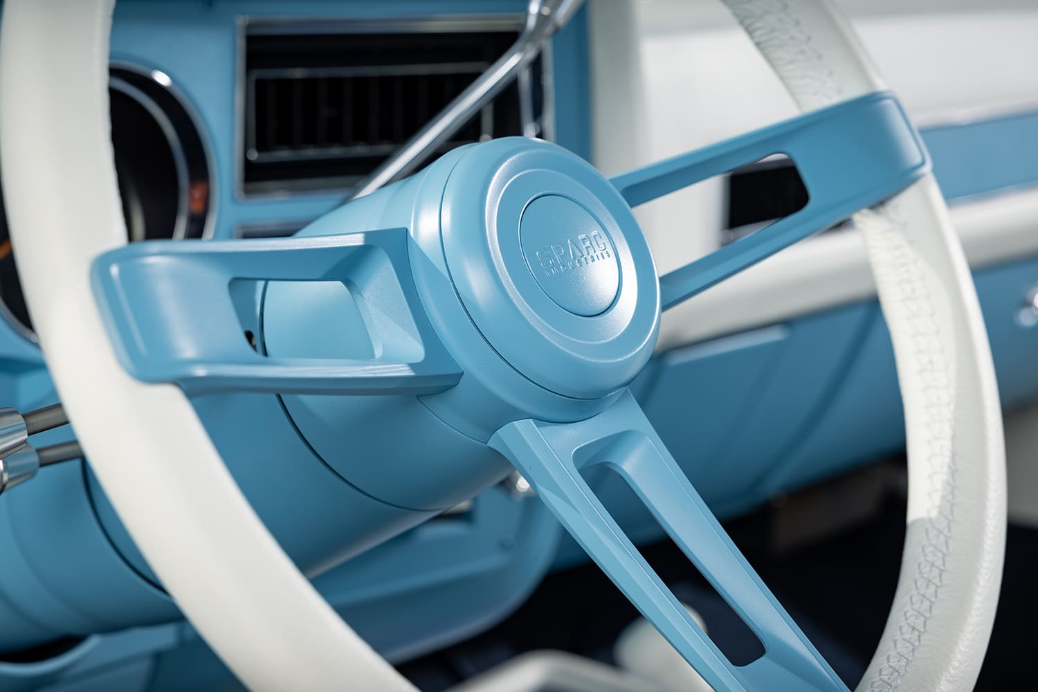 Close-up detail of a custom Sparc Industries three-spoke steering wheel featuring a light blue painted billet center and horn cap with a white leather-wrapped rim inside a 1976 Chevy truck.