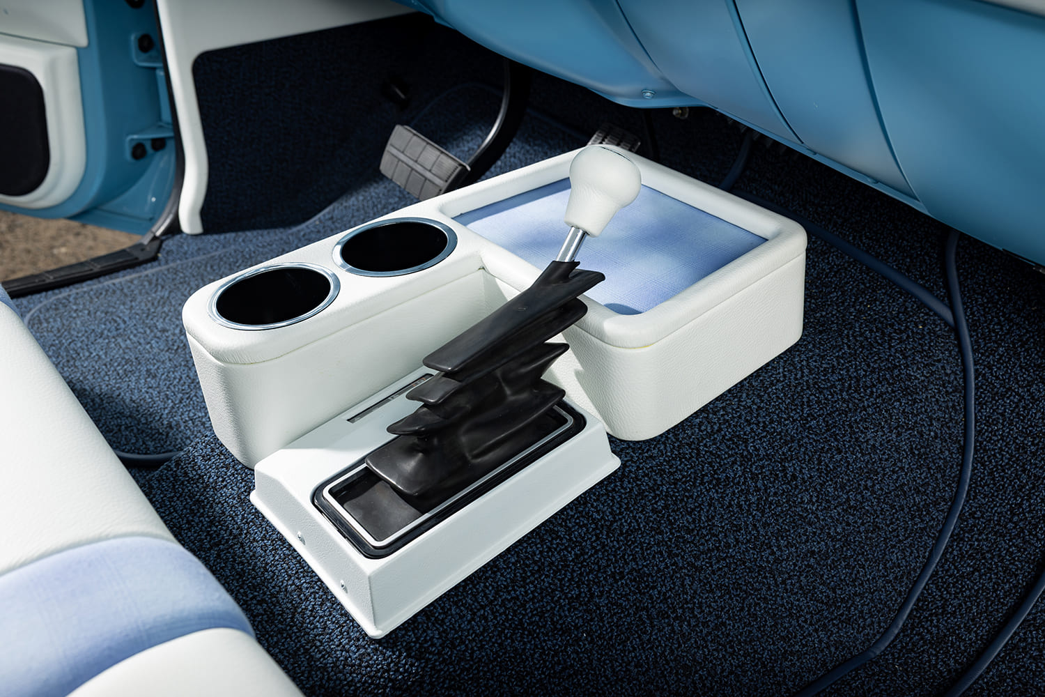 Custom white upholstered floor console featuring dual cup holders, storage tray, and an automatic floor shifter over dark blue loop carpet in a 1976 Chevy truck.