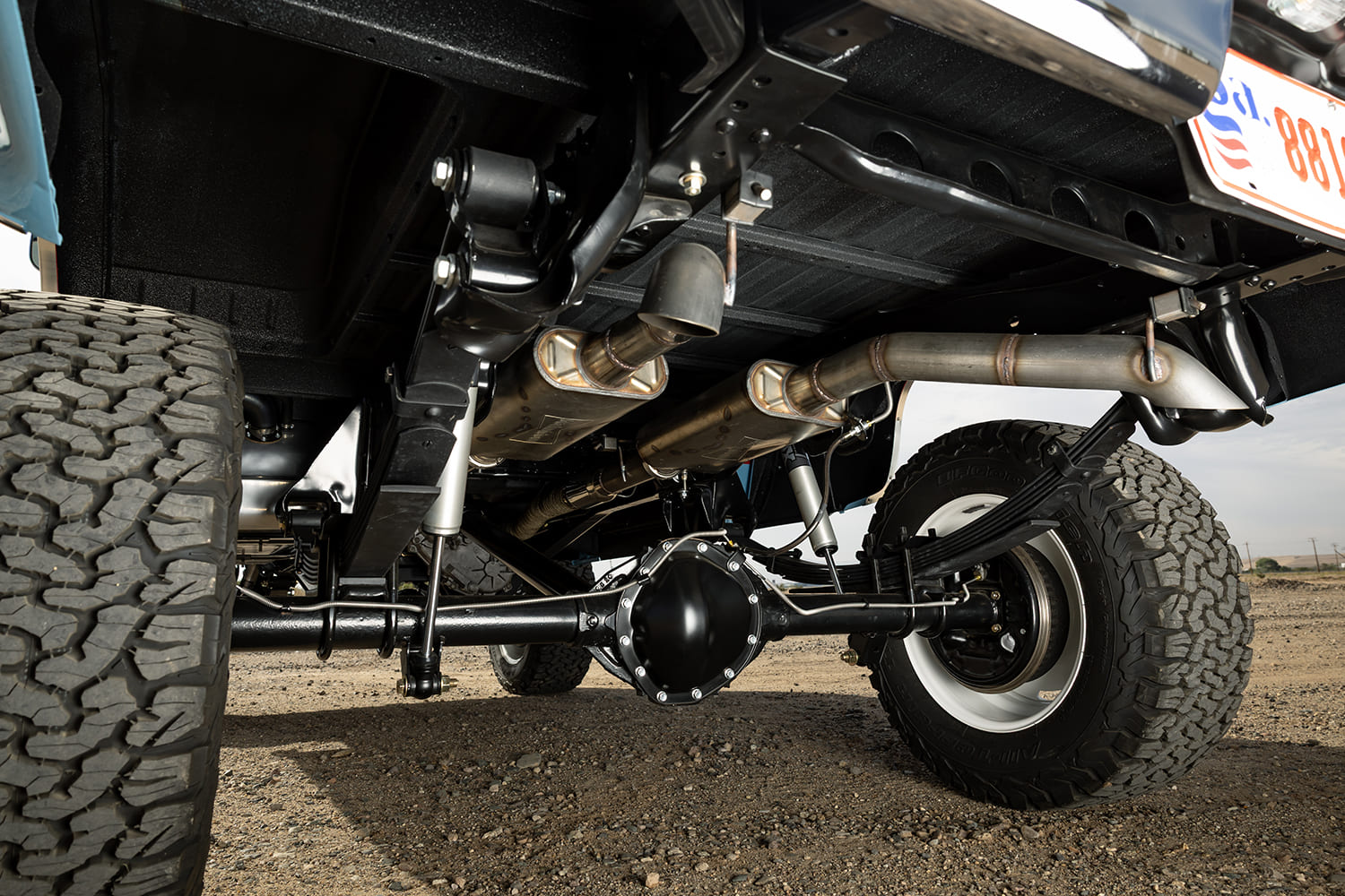 Detailed undercarriage view of a lifted 1976 Chevy 4x4 pickup truck showing the rear axle, leaf springs, Fox shocks, and custom stainless steel dual exhaust system with turndowns.