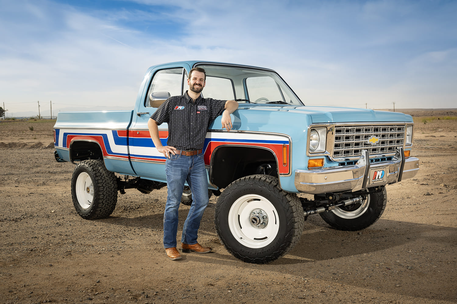 rockford smith leaning against a restored light blue 1976 Chevy 4x4 pickup truck featuring red and blue retro body stripes, lifted suspension, and white wheels in an outdoor desert setting.