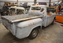 Rear three-quarter view of a vintage patina 1955-1959 Chevrolet Task Force Stepside pickup truck undergoing restoration in a garage workshop with a cat looking out the rear window.