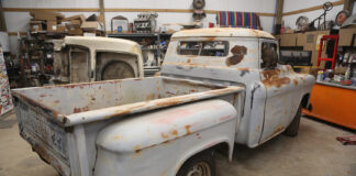 Rear three-quarter view of a vintage patina 1955-1959 Chevrolet Task Force Stepside pickup truck undergoing restoration in a garage workshop with a cat looking out the rear window.