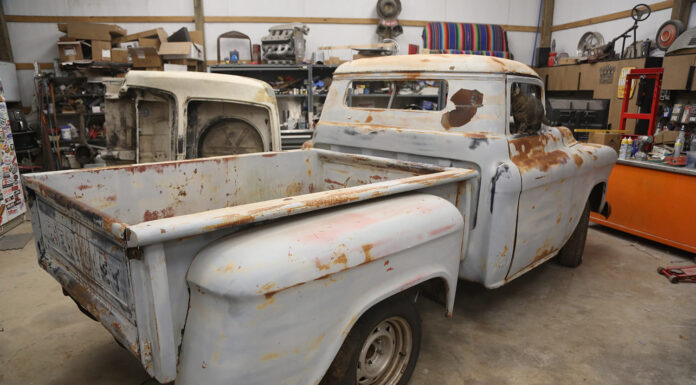 Rear three-quarter view of a vintage patina 1955-1959 Chevrolet Task Force Stepside pickup truck undergoing restoration in a garage workshop with a cat looking out the rear window.