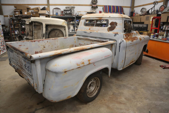 Rear three-quarter view of a vintage patina 1955-1959 Chevrolet Task Force Stepside pickup truck undergoing restoration in a garage workshop with a cat looking out the rear window.