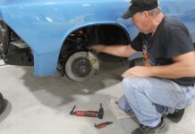 Mechanic inspecting the front disc brake caliper, rotor, and suspension assembly on a classic blue car supported by jack stands.