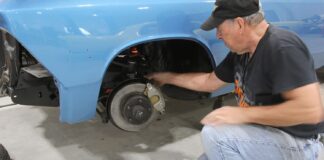 Mechanic inspecting the front disc brake caliper, rotor, and suspension assembly on a classic blue car supported by jack stands.
