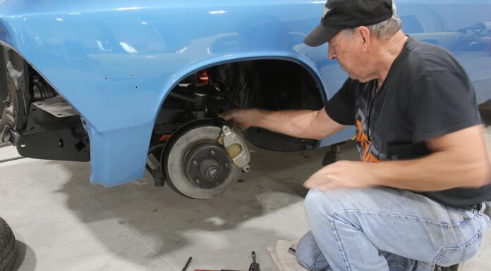 Brake Maintenance Tips – Keep Your Muscle Car Stopping Properly Mechanic inspecting the front disc brake caliper, rotor, and suspension assembly on a classic blue car supported by jack stands.
