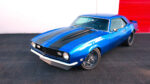 High-angle front three-quarter view of a bright blue 1968 Chevrolet Camaro SS restomod with black cowl stripes and custom gray wheels.