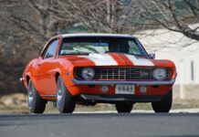 Low-angle front shot of an original Hugger Orange 1969 Chevrolet Camaro Z/28 with white racing stripes, cowl induction hood, and USA-1 front novelty plate on asphalt.