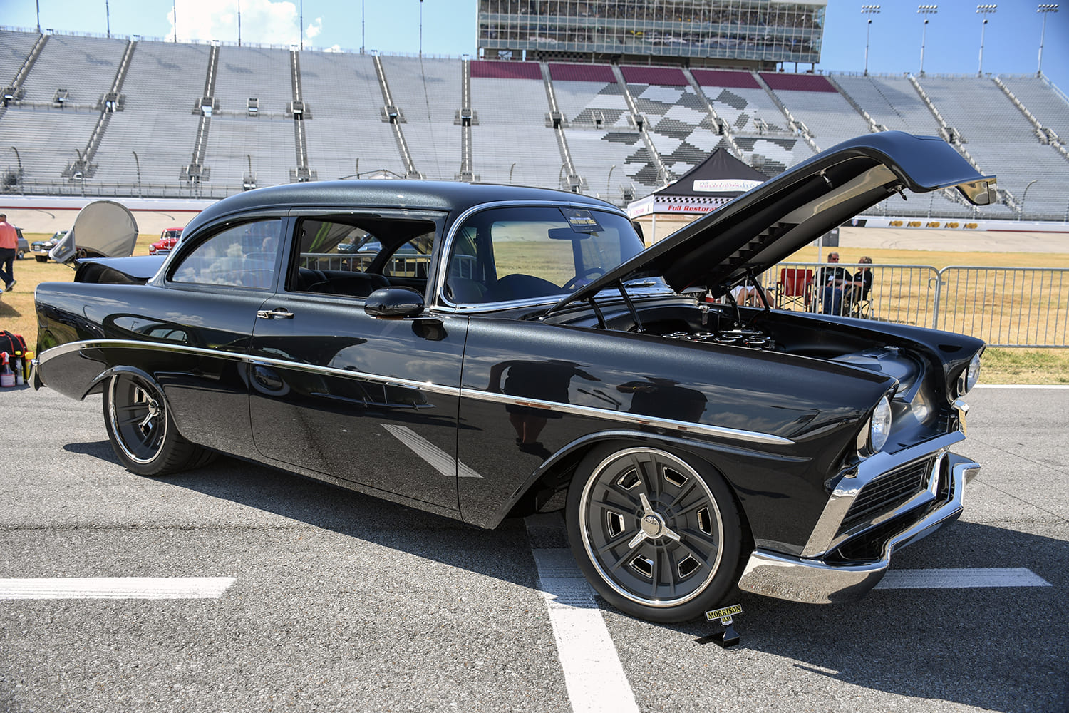 Black pro-touring 1956 Chevrolet two-door sedan with an open hood, and custom gray alloy wheels on Nashville speedway asphalt apron.