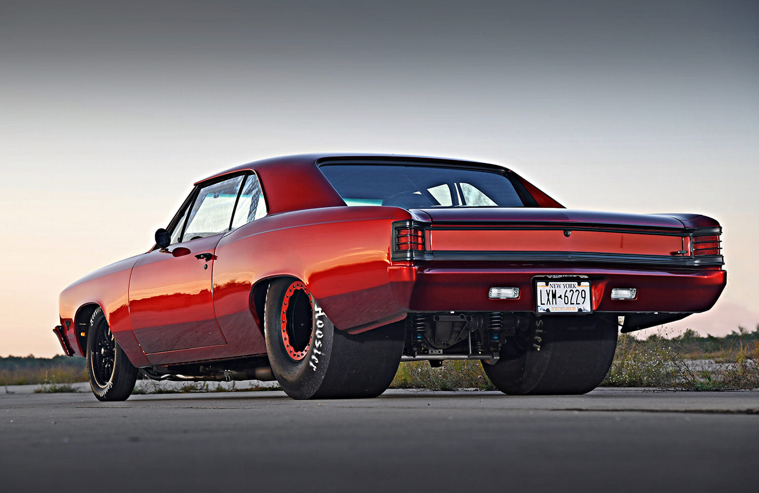 Ground-level rear three-quarter view of a tubbed 1967 Chevelle in metallic red paint highlighting wide Hoosier racing slicks and rear suspension.