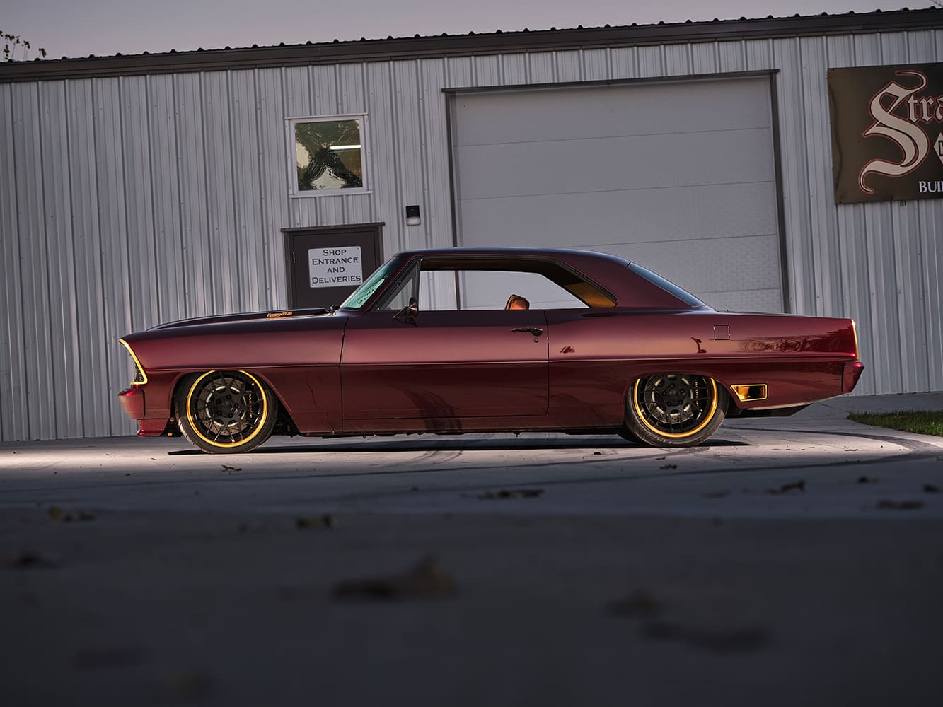 Low-slung side profile of a pro-touring 1967 Chevrolet Chevy II Nova coupe with deep candy red paint, side exhaust outlets, and gold-rimmed performance wheels.