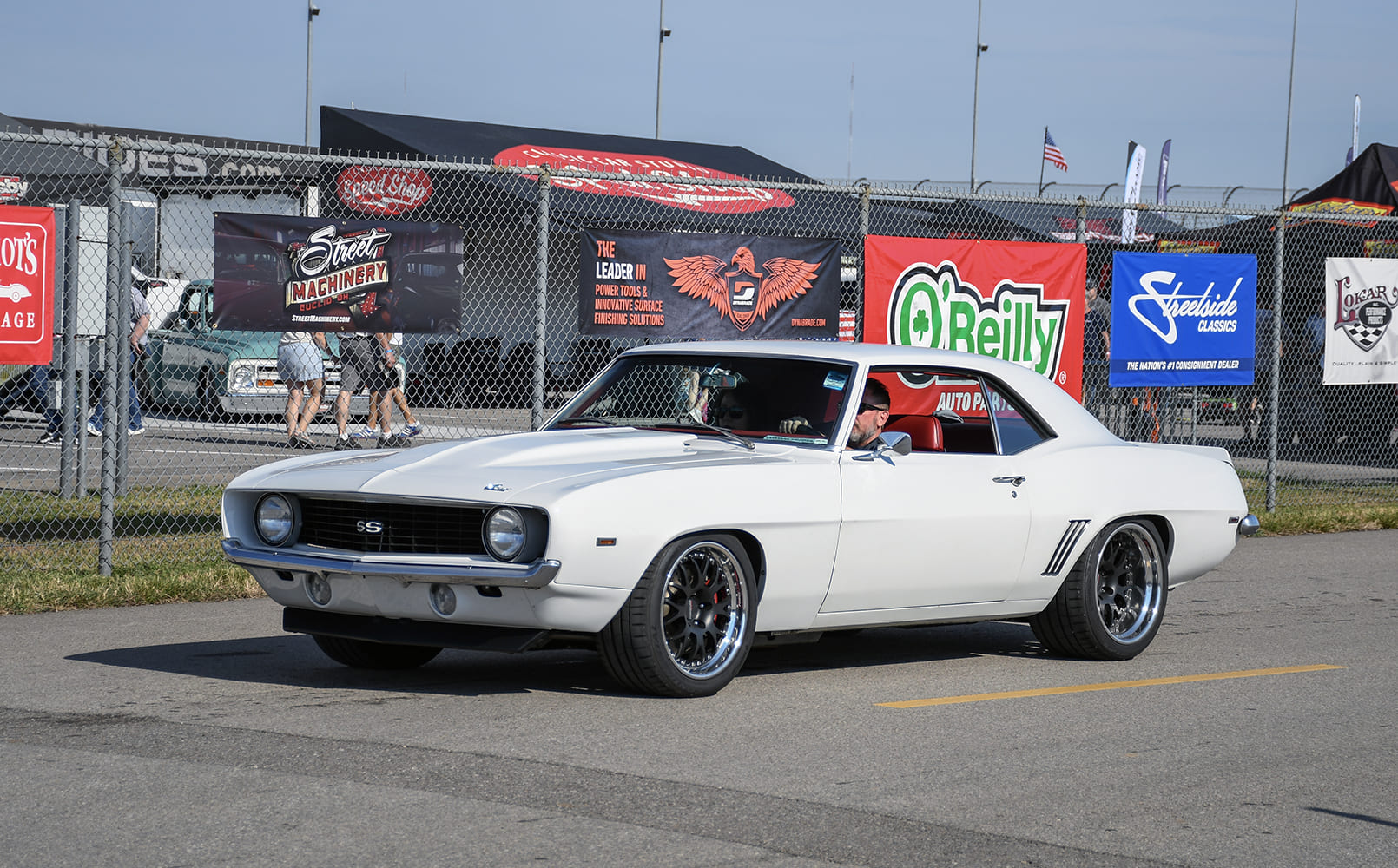Front three-quarter rolling shot of a white 1969 Chevrolet Camaro SS coupe equipped with black multi-piece wheels driving past outdoor vendor tents.