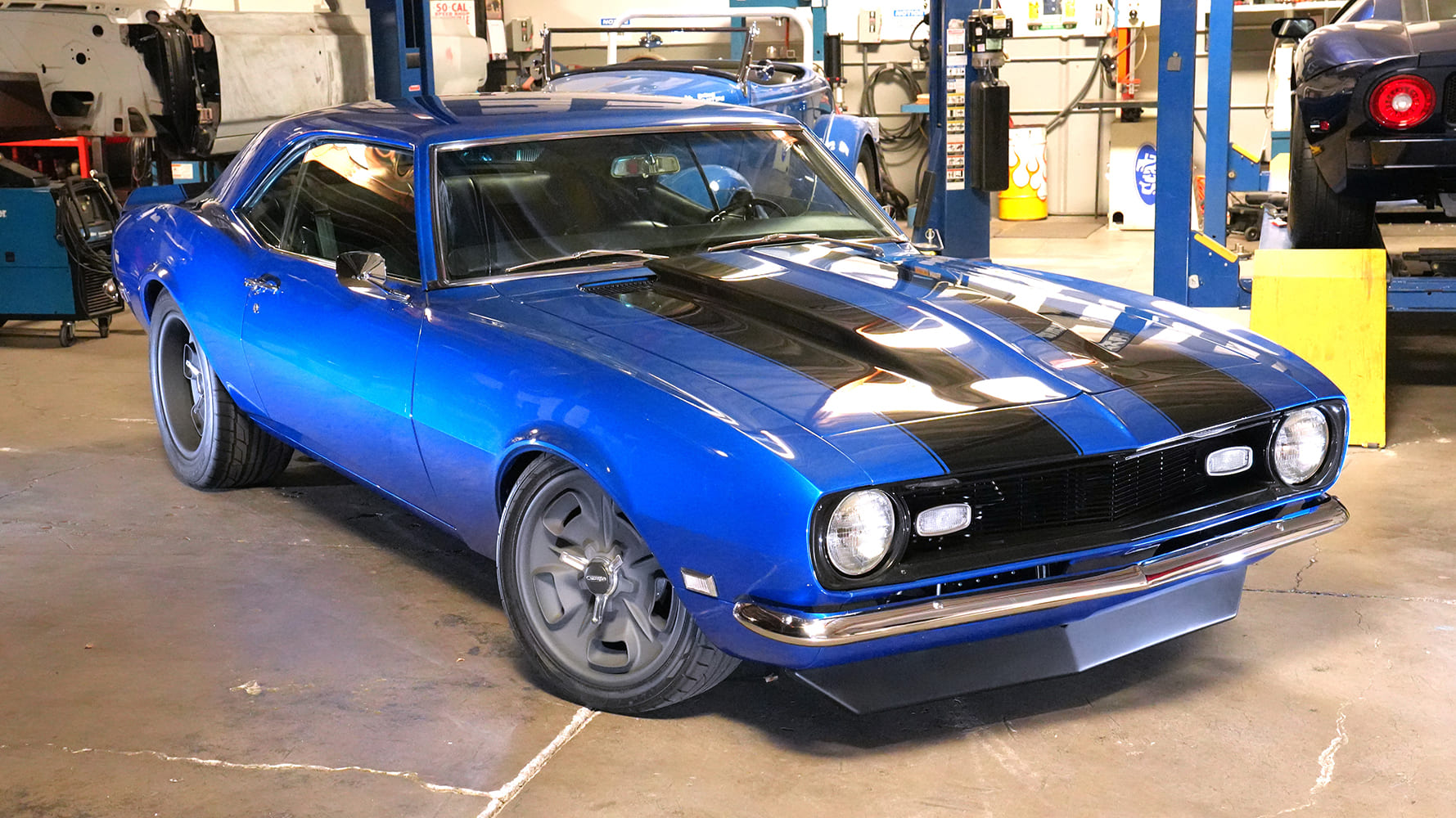 Low-angle front profile showing the lowered pro-touring stance, front air dam, and tucked multi-spoke wheels on a blue 1968 Camaro.