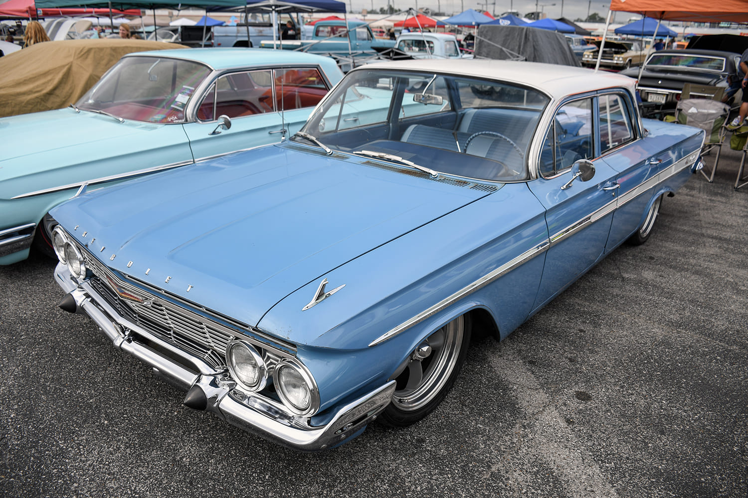 Elevated front-end view of a lowered powder-blue 1961 Chevrolet four-door sedan with a white roof, polished chrome grille, and custom wheels at a car show.