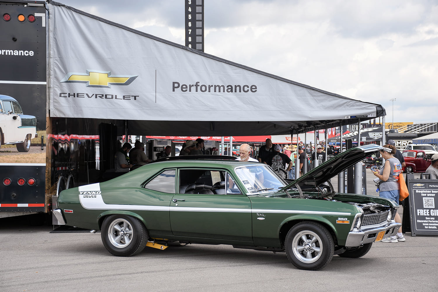 Side profile of a forest green Yenko Deuce Nova with white side stripes, open hood, and vintage Torq Thrust wheels displayed beside a Chevrolet Performance trailer.