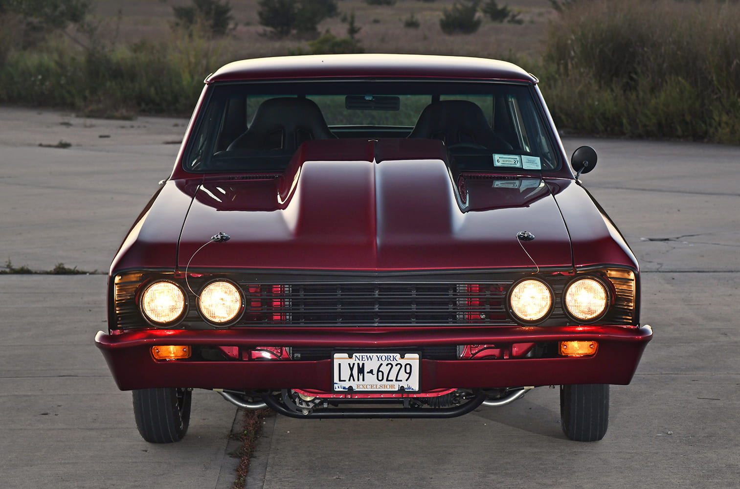 Head-on shot of a dark red metallic 1967 Chevelle street-strip car with illuminated quad headlights, black billet grille, and tall cowl-induction hood.