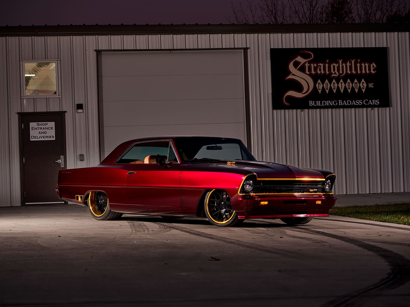 Deep candy red 1967 Chevy Nova pro-touring restomod parked in front of the Straightline Customs shop at dusk.
