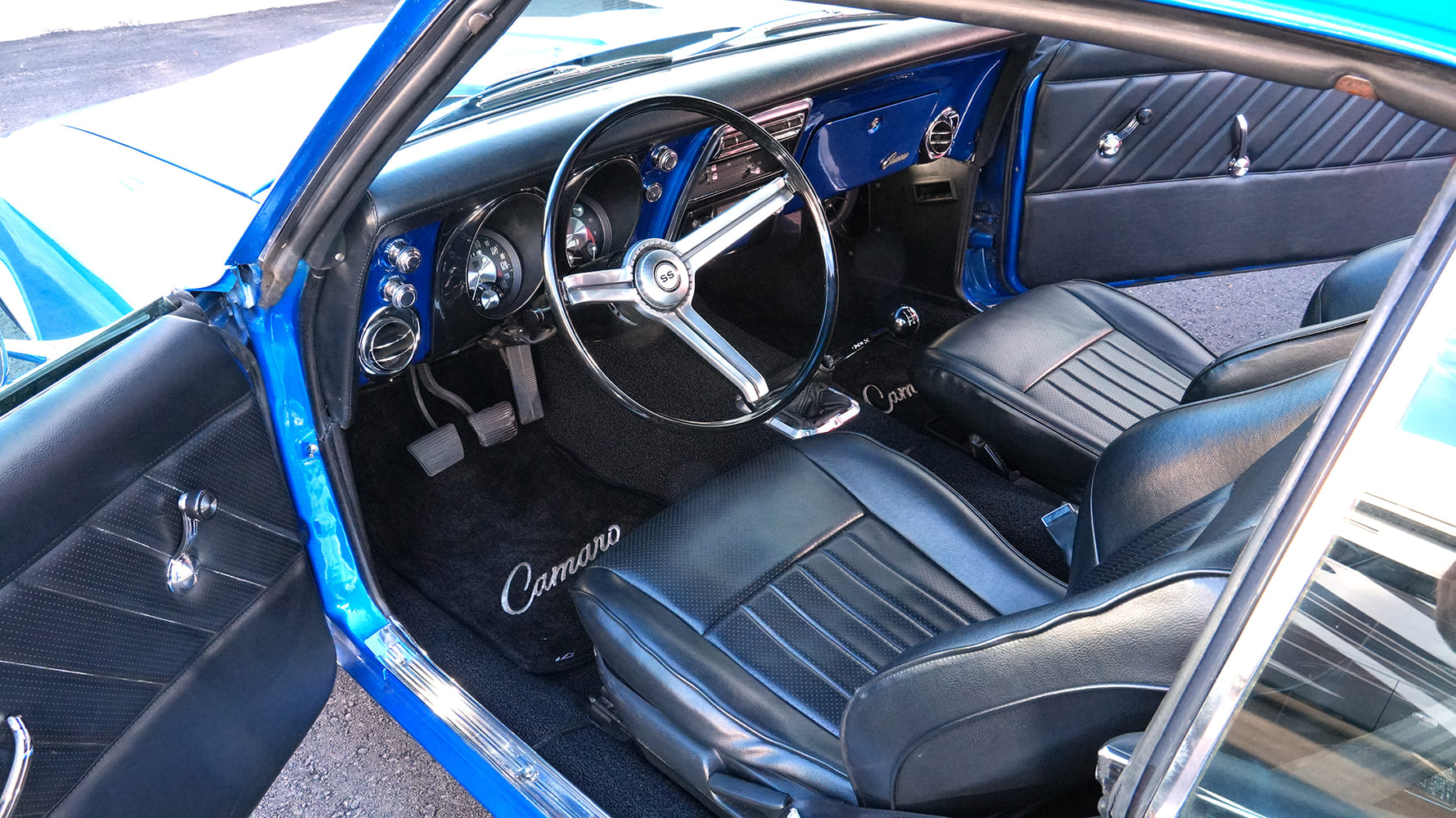Open-door interior view of a 1968 Chevy Camaro showcasing black leather sport seats, color-matched blue dash, floor shifter, and SS steering wheel.