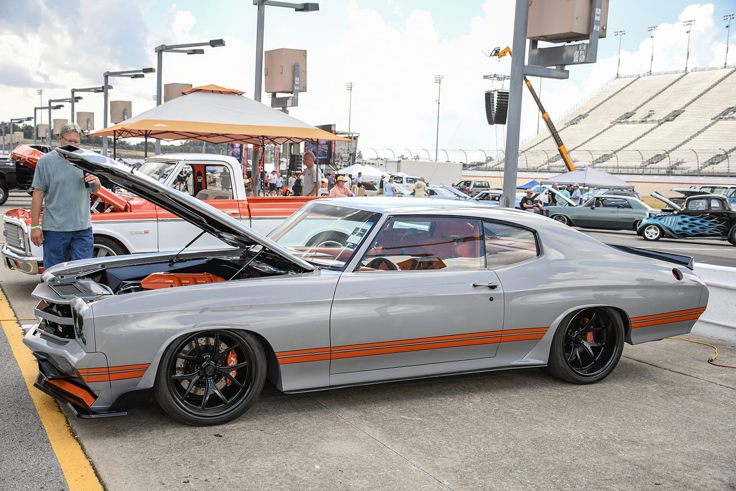 Low-profile side view of a modern gray 1970 Chevrolet Chevelle restomod featuring orange side stripes, flared fenders, black wheels, and an exposed engine bay.