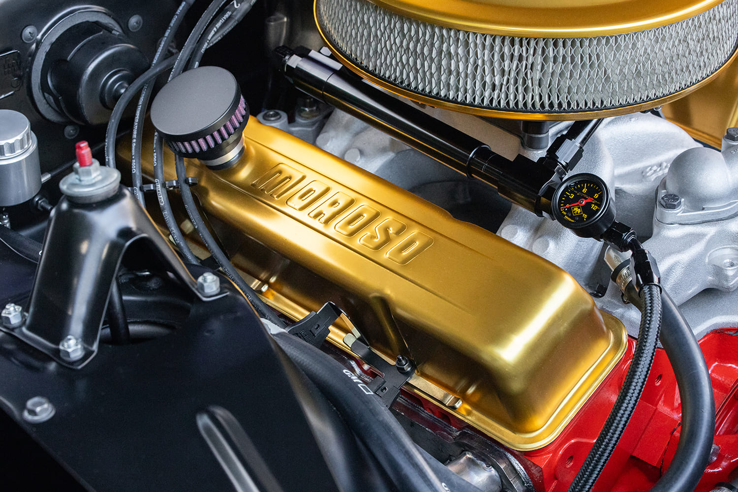 Close-up of gold-stamped Moroso valve covers, breather cap, braided black fuel lines, and inline fuel pressure gauge on a vintage Chevy V8.