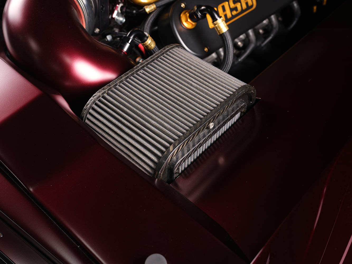 Custom carbon-fiber framed high-flow air filter connected to a candy red intake tube in a pro-touring Chevy Nova engine bay.