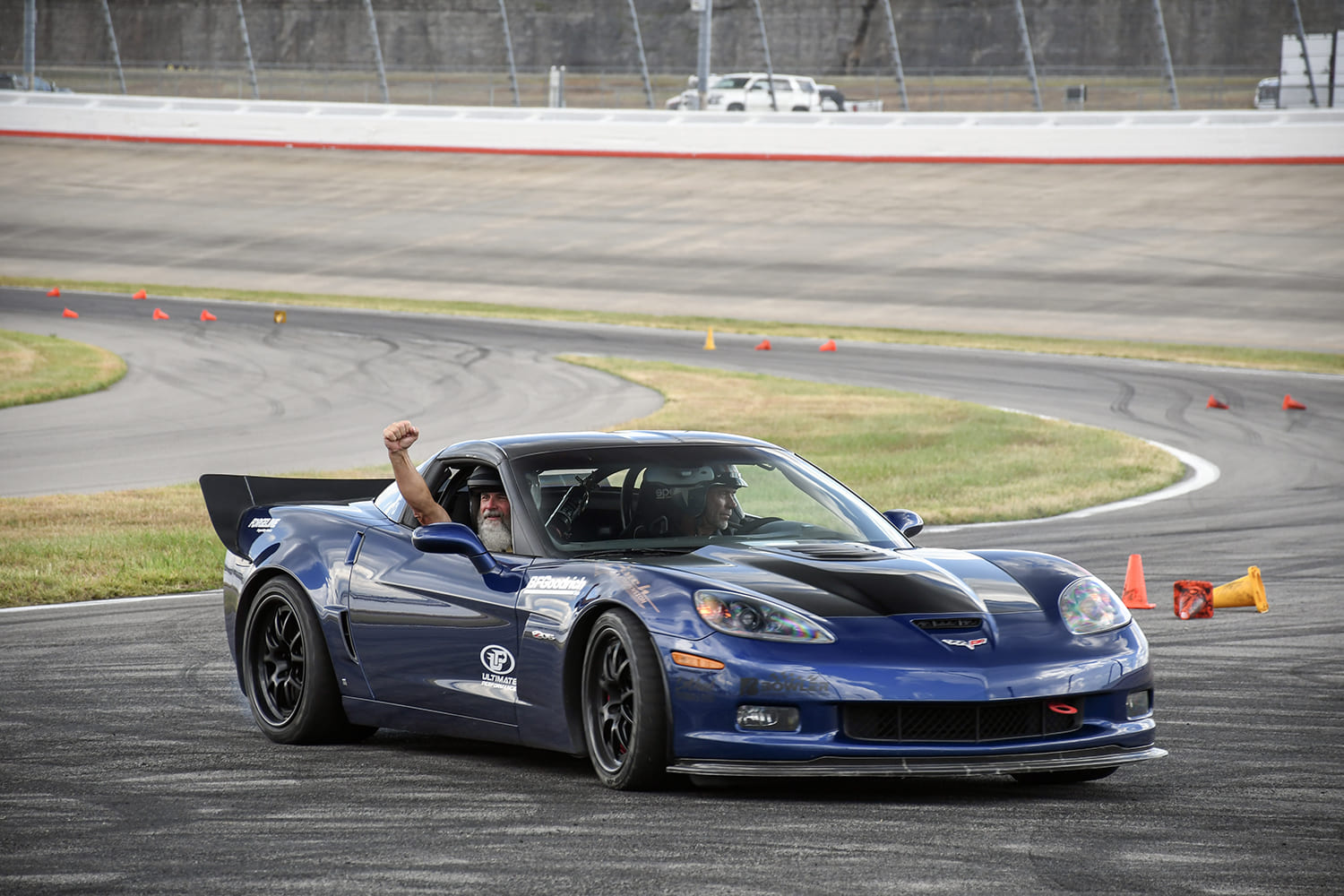 Blue C6 Chevrolet Corvette Z06 turning around orange cones on an autocross track with the driver raising a fist in celebration through the open window.