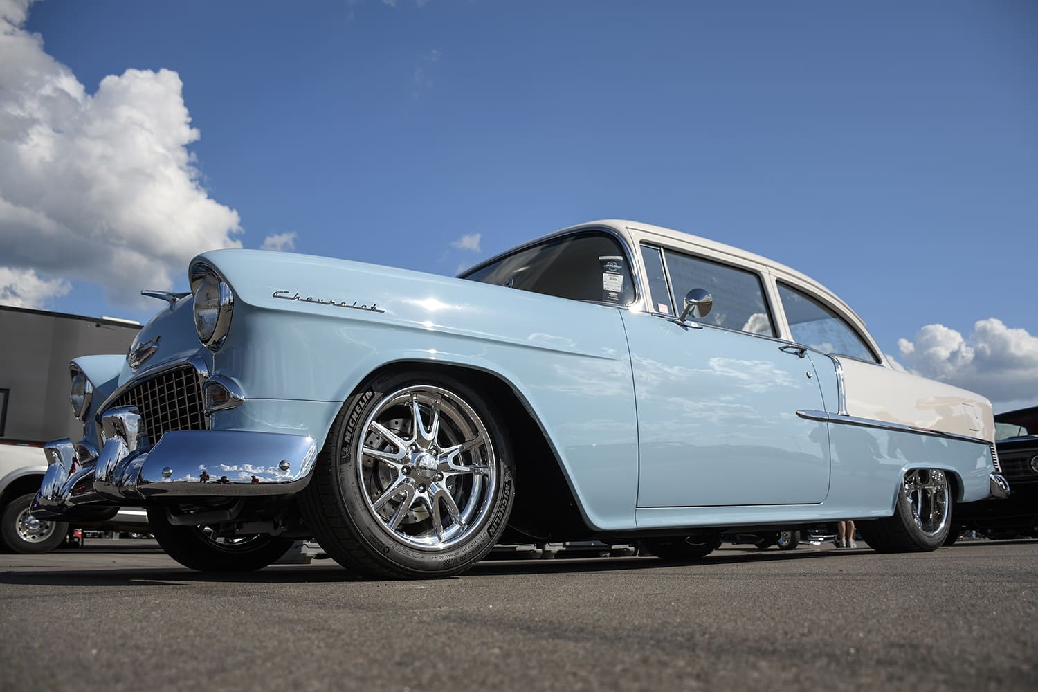Low-angle front three-quarter perspective of a two-tone baby blue and cream 1955 Chevrolet Bel Air showing chrome five-spoke wheels under a bright blue sky.