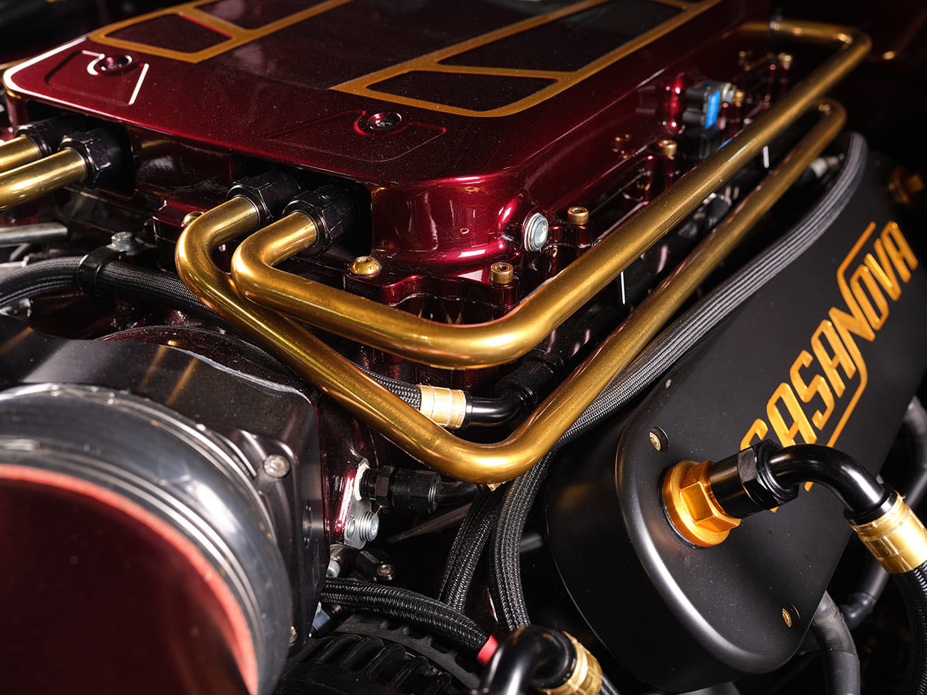 Close-up shot of the custom gold hard plumbing lines, candy red supercharger housing, and black Casanova valve cover on a high-performance restomod engine.
