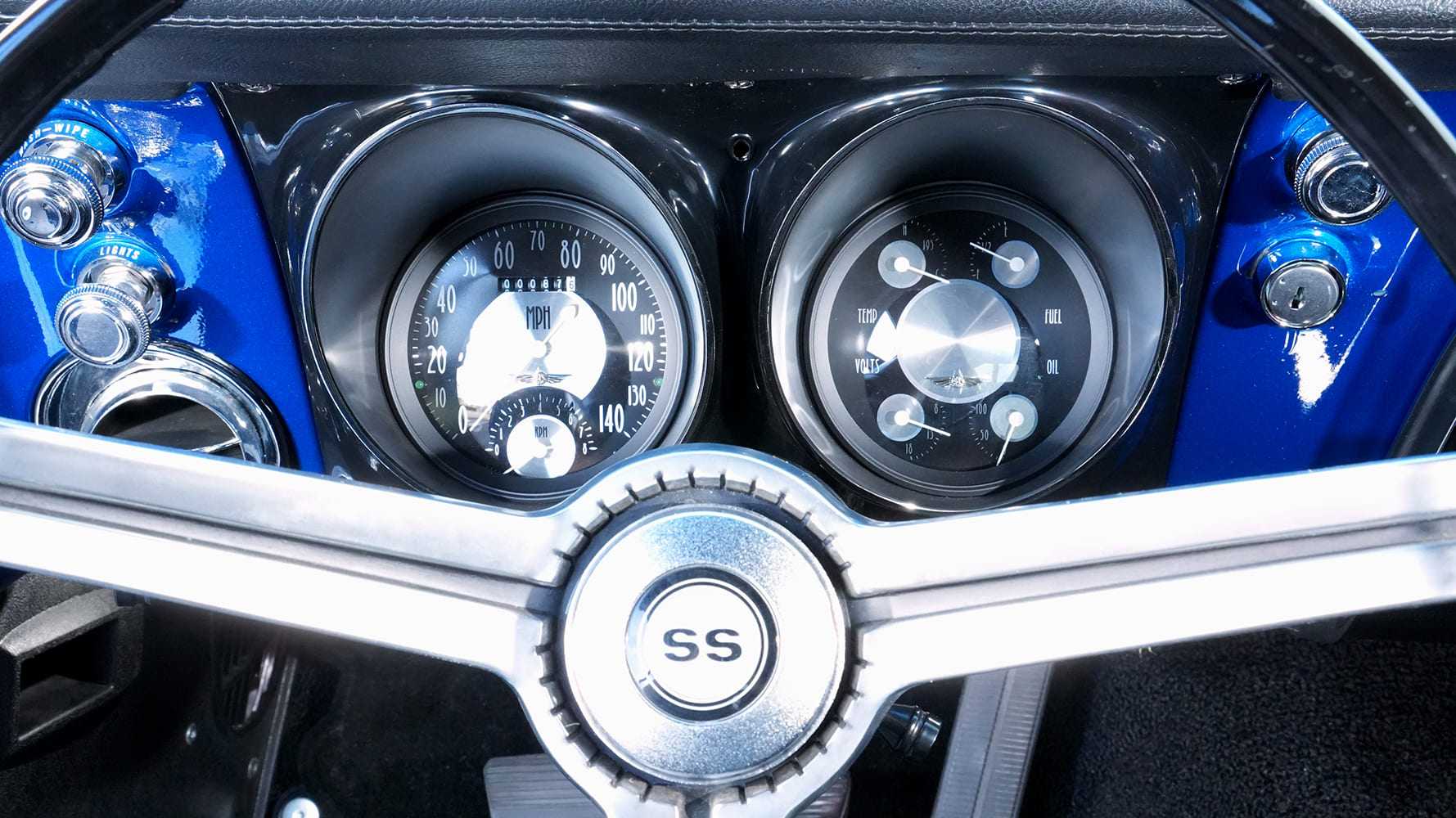 Driver perspective of the three-spoke SS steering wheel and retro-styled modern analog gauge cluster set in a blue painted dashboard.