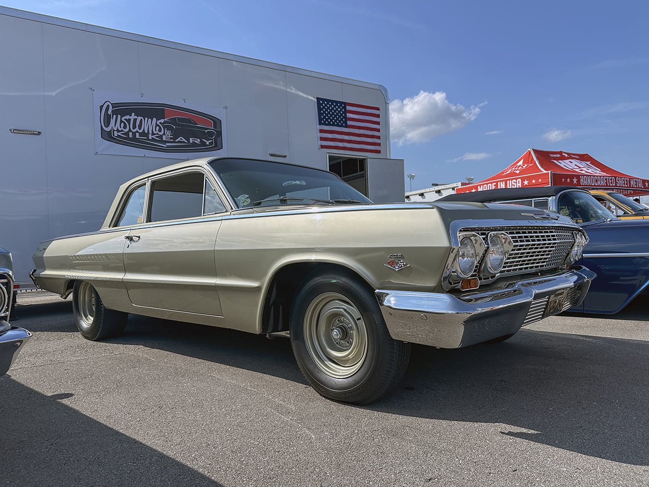 Front three-quarter angle of a champagne gold 1963 Chevrolet Impala post coupe fitted with color-matched steel wheels parked outside a Kilkeary race transporter.