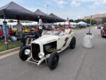 Front three-quarter view of an off-white 1932 Ford roadster hot rod with "Banger Power" hood lettering carrying passengers down the fairgrounds thoroughfare past spectator canopies.